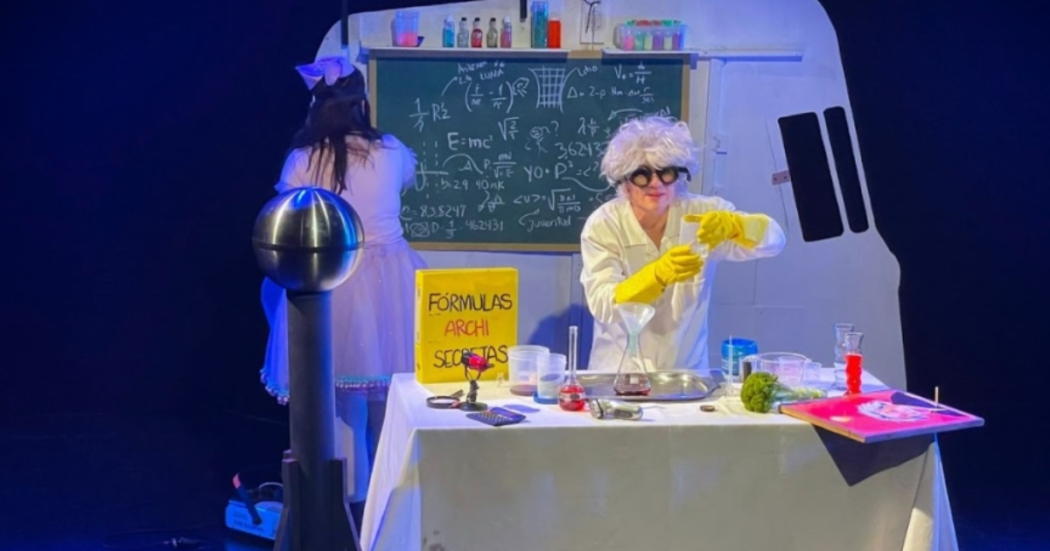 Imagen de un científico que prepara sus formulas en una mesa, a su respaldo en un tablero hay una mujer que escribe en un tablero