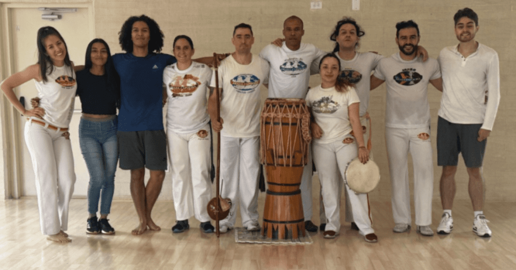 Imagen de seis bailarines de capoeira en escenario acompañados de instrumentos músicales