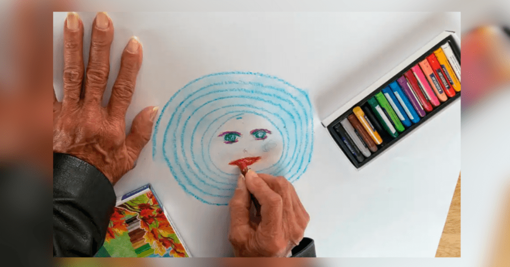 Imagen de una mano de una persona mayor que dibuja una cara con circulos de color azul.