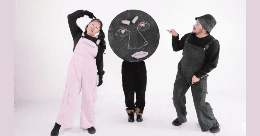 Imagen de tres personajes, uno viste un overol rosado y el otro verde oscuro, el tercer personaje lleva un gran círculo negro con un rostro dibujado en tiza que cubre su cuerpo.