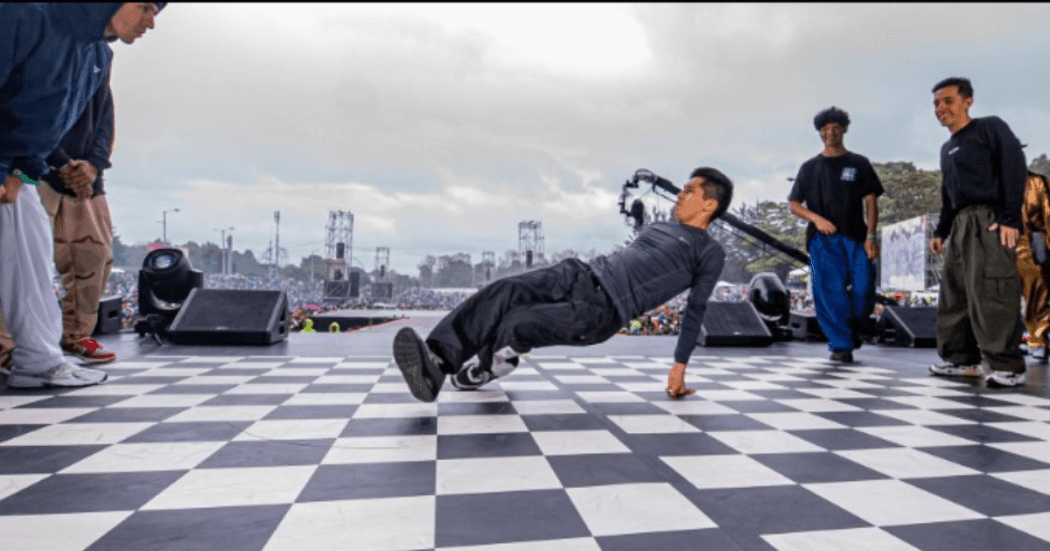 Grupo de jóvenes bailando en una pista con apariencia de ajedrez, de fondo se ve mucha gente observándolos