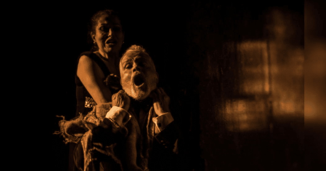 Imagen de la puesta en escena donde se ve a una mujer que ahorca por la espalda a un hombre de barba que está sendado en un silla