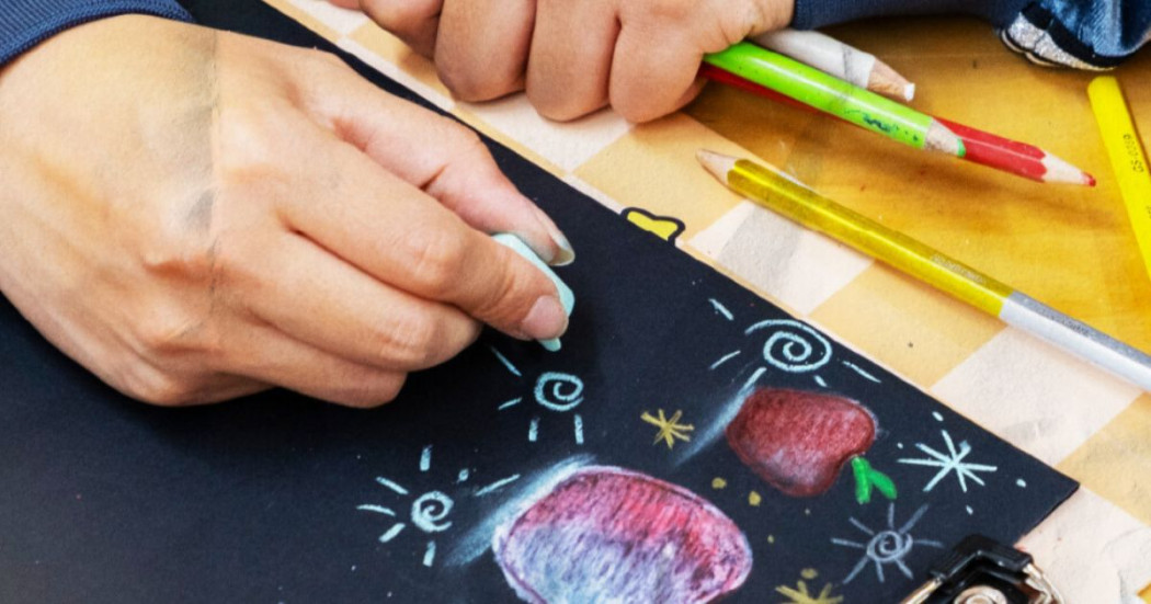 Imagen de una mano con una tiza sobre cartelera negra con dibujos y colores