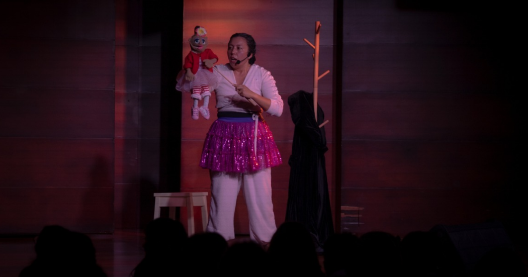 Imagen de los protagonistas de la obra: una mujer joven vestida de balnco con un delantal y un títere femenino con vestido rojo.