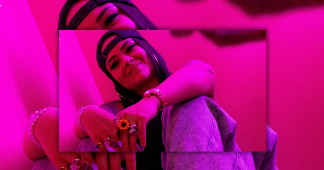 Imagen de " Angelik DK" sentada con gorra y anillos en un fondo rosado