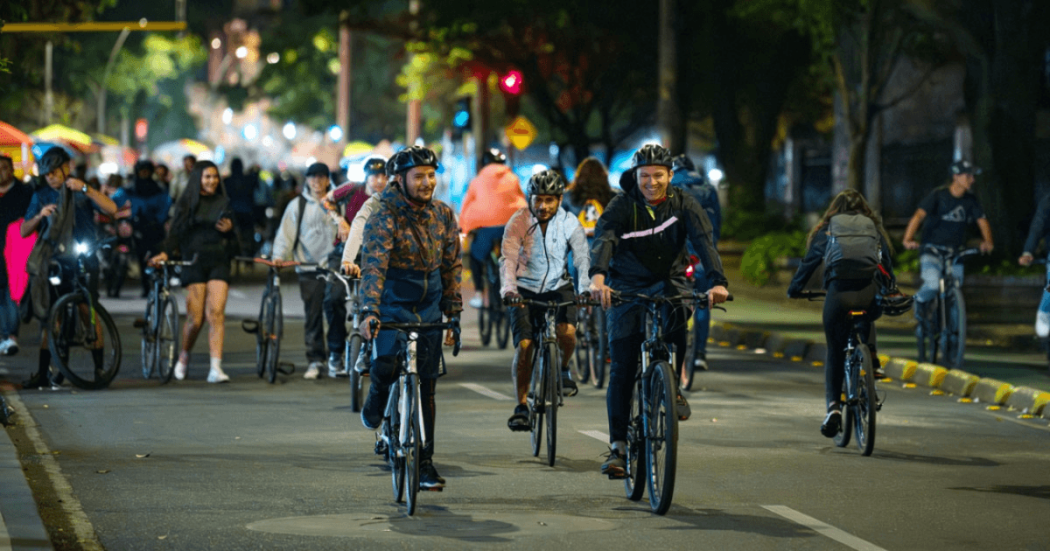 Ciclistas y peatones recorren una avenida de Bogotá durante la Ciclovía Nocturna, con grupos de personas en bicicleta, alumbrado público, árboles y actividad recreativa en horario nocturno.