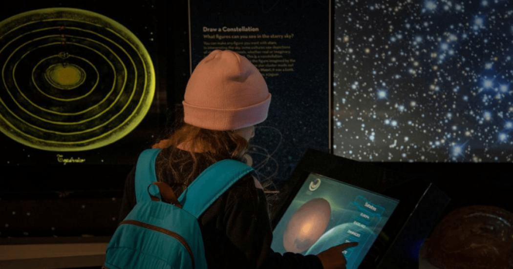 Imagen de una niña con maleta y gorro de espaldas interactuando con pantalla en el Planetario de Bogotá
