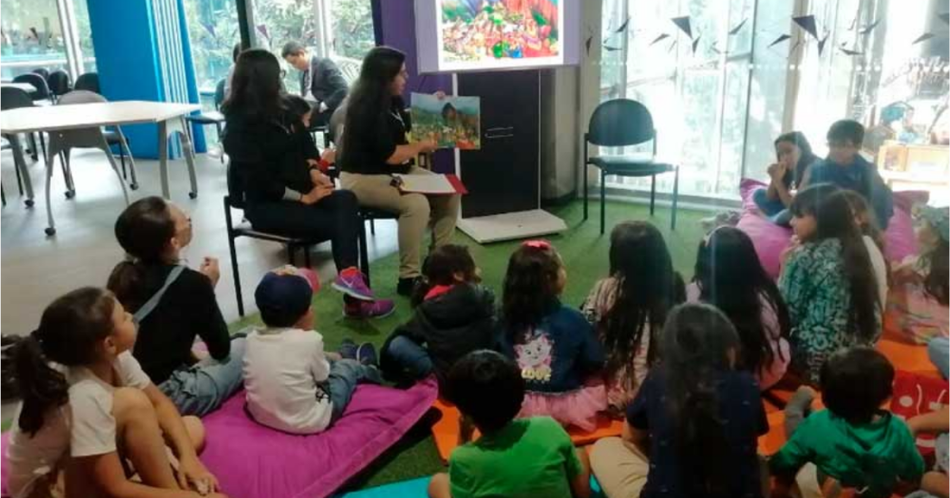 Iamgen de varios niños y niñas sentados en colchonetas atendiendo la charla de la tallerista que indica en un libro.