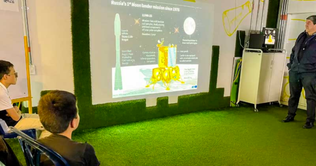Imagen de dos niños sentados observando una presentación en una pantalla gigante, al lado hay un hombre adulto hablando. 