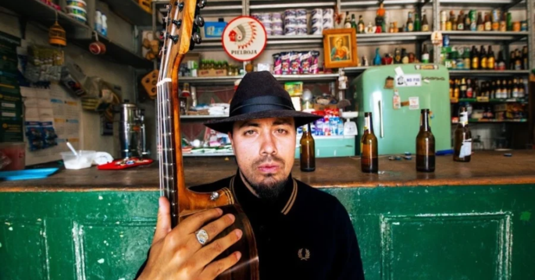 Imagen del cantante en una tienda de barrio con una guitarra en la mano, de fondo se ven varias botellas de cerveza.