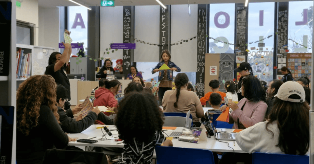 Actividad cultural en una biblioteca pública de Bogotá, con varias personas sentadas alrededor de mesas mientras observan una presentación en un escenario al frente. En el espacio se ven estanterías, elementos decorativos y letras colgantes.