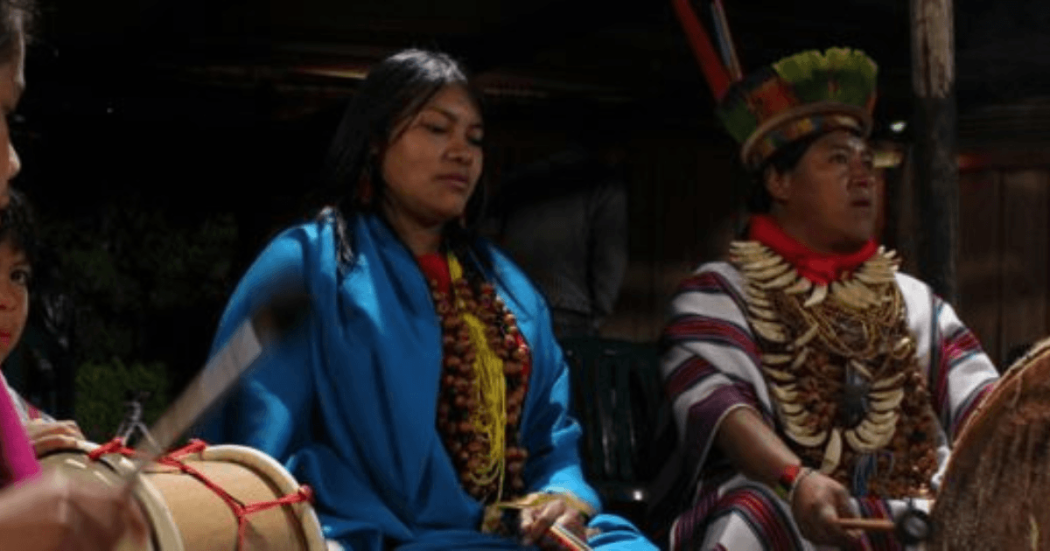 Imagen de personas indigenas sentadas con corona de plumas y manta azul