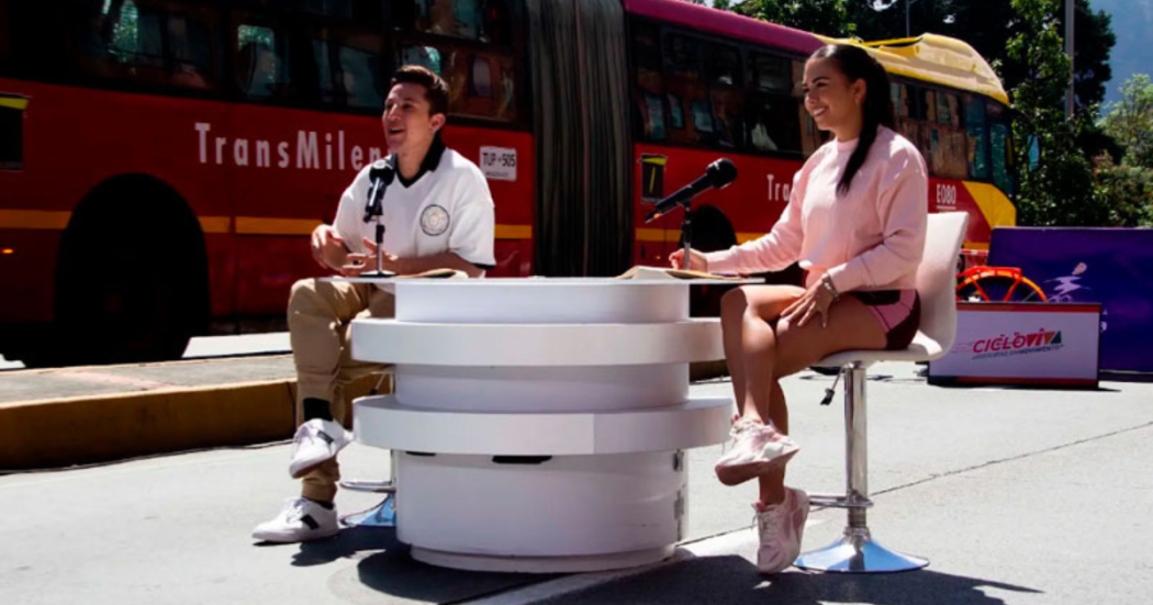 Imagen de un hombre y una mujer, presentadores de Cicloviva sentados en una vía de Bogotá, de fondo se ve un bus de Transmilenio rojo.