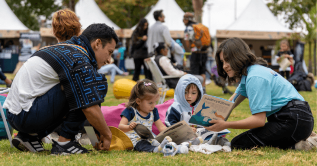 Imagen de dos adultos con niños en un parque leyendo un libro 