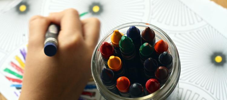 Las manos de un niños coloreando con crayolas de colores, al lado hay un vaso lleno de estas crayolas 