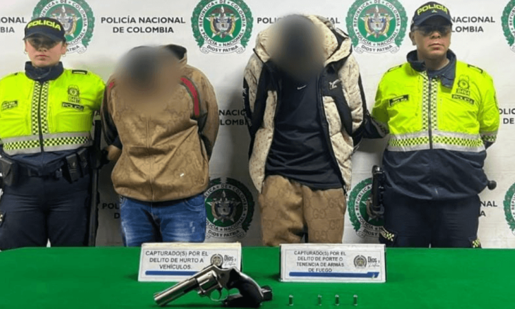 Dos capturados en medio de dos Policías. Sobre una mesa está el arma y los car