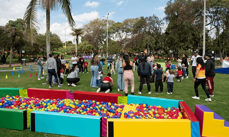 Personas adultas y menores participan en actividades recreativas sobre una zona verde de parque; en primer plano hay piscinas secas con pelotas de colores y al fondo se observan conos, árboles y grupos reunidos al aire libre.