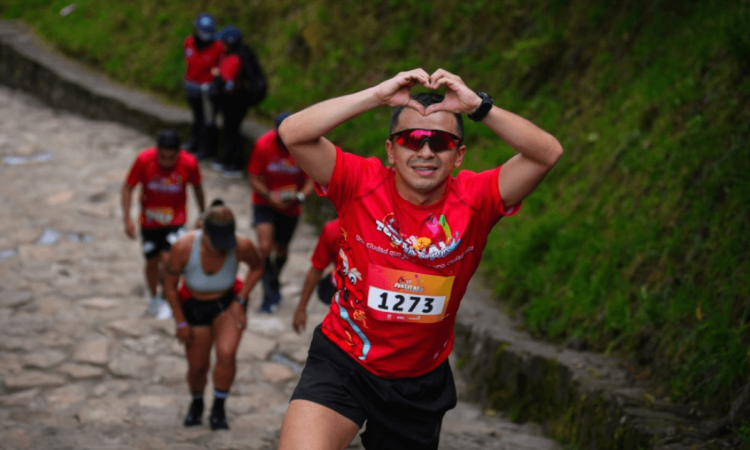 Participantes ascienden por el sendero de Monserrate, en primer plano un corredor con el número 1273 realiza un gesto de corazón con las manos mientras continúa el recorrido.