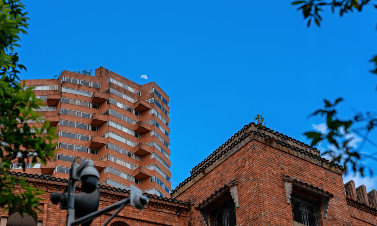 Edificaciones de ladrillo y torre de apartamentos en Bogotá, con vegetación en primer plano y cielo azul al fondo, en una escena urbana residencial.