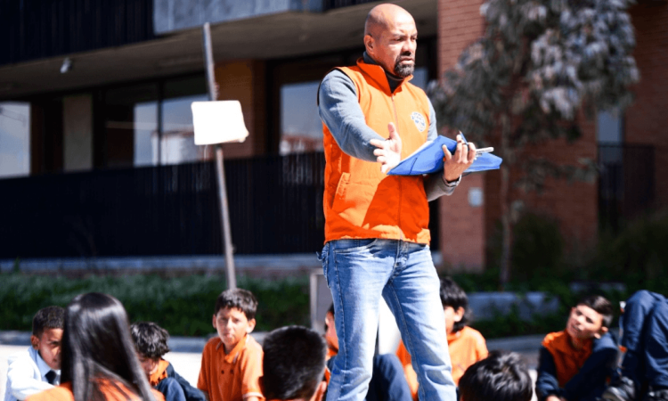 Foto que muestra un hombre con una carpeta en las manos y estudiantes sentados en el piso 