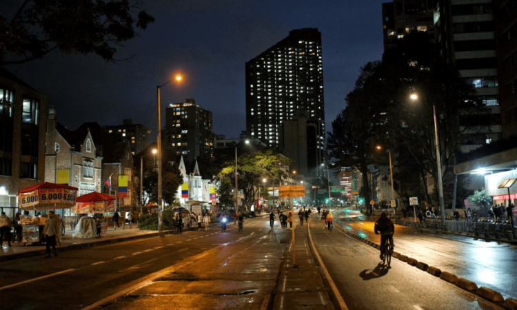 Ciclistas y peatones recorren una avenida iluminada durante la Ciclovía Nocturna de Bogotá, con edificios, comercio, alumbrado público y pavimento húmedo visibles en horario nocturno.