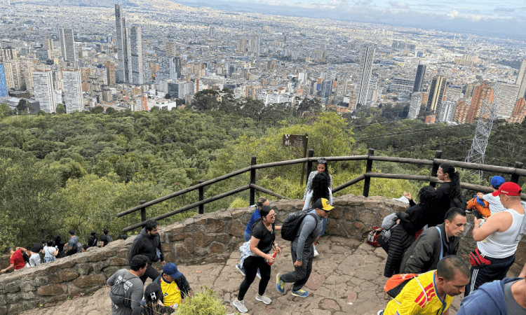 Personas acienden por el sendero de Monserrate con un fondo panorámico de la ciudad de Bogotá donde se observan edificios y zonas residenciales