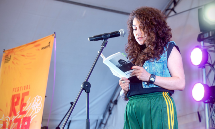 Imagen de una mujer joven en una tarima leyendo un libro de poesia, al lado inzquierdo se ve una pancarta del concurso Reverso.