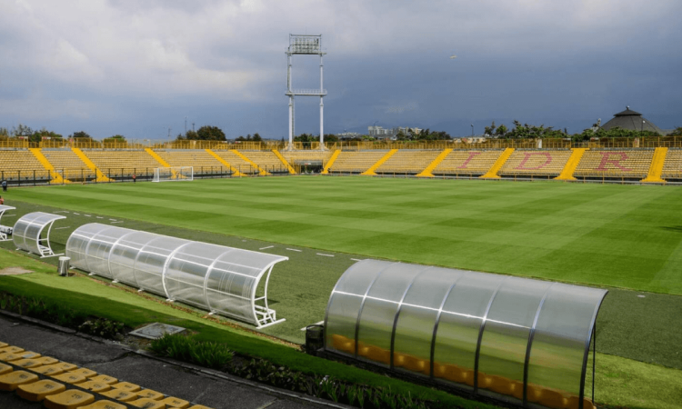 Estadio de Techo de Bogotá, con una cancha de fútbol de césped natural rodeada por graderías amarillas, bancas técnicas cubiertas y un cielo nublado al fondo.