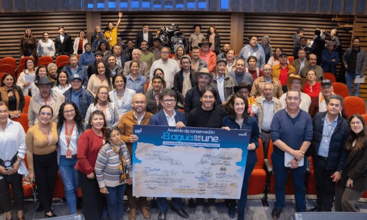 Grupo de personas reunidas en un auditorio con sillas rojas. En el centro de la imagen varias personas sostienen un tablero gigante con el texto: “Acuerdo de conservación. El agua que nos une”, junto a múltiples firmas