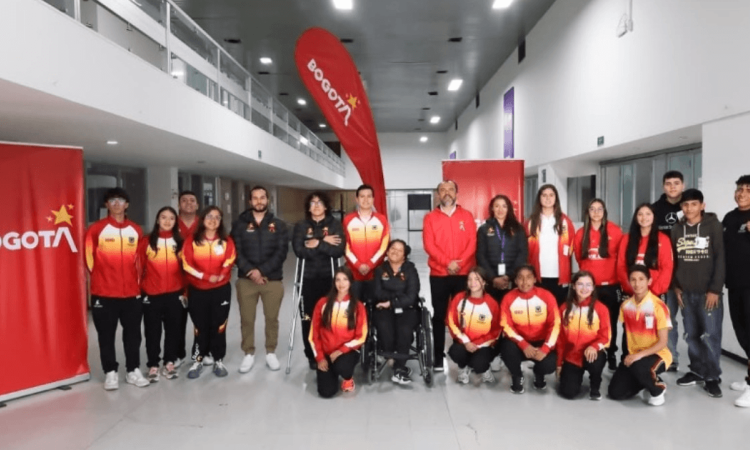 Grupo de deportistas del Equipo Bogotá, junto a personal de apoyo, posan para una fotografía en el hall interior de un edificio. Varias personas visten chaquetas deportivas en tonos rojo, amarillo y blanco.