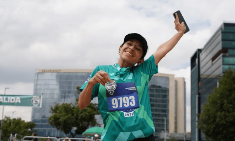 Participante de la Bimbo Global Race con camiseta verde y dorsal 9193 levanta un brazo mientras corre. Al fondo se observan edificaciones y el cielo despejado.
