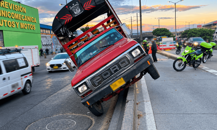 Camión rojo inclinado sobre el separador de una vía de Bogotá, con dos ruedas elevadas. A un costado hay motocicletas de tránsito, personal en la calzada y vehículos circulando en varios carriles.