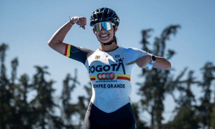 Gabriel Rueda, patinadora bogotana, posa con uniforme de la Liga de Patinaje de Bogotá y casco deportivo en una pista al aire libre, señalando el logo de Bogotá en la camiseta, con árboles desenfocados al fondo y cielo despejado.