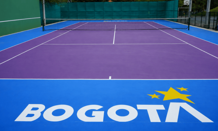 Cancha de tenis con superficie morada y zonas laterales azules, delimitada por líneas blancas y una red en el centro. En el primer plano se observa el logo de Bogotá en color blanco y amarillo sobre la superficie de la cancha.