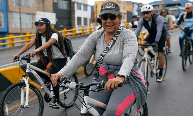 Personas en bicicleta recorren una vía urbana durante una jornada de Ciclovía. En primer plano, una persona vestida con ropa deportiva gris y gorra conduce una bicicleta blanca