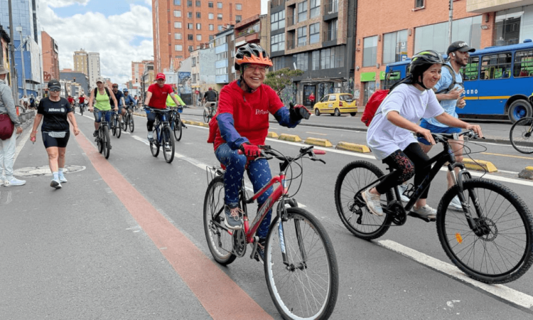 Ciclistas y peatones transitan por una vía urbana durante una jornada recreativa. En primer plano, dos personas montan bicicleta con casco mientras otras personas avanzan en bicicleta y caminando por la calzada.