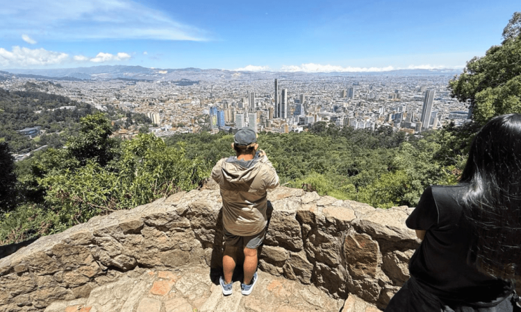 Vista panorámica de Bogotá desde un mirador en Monserrate. En primer plano se observa un muro de piedra y dos visitantes de espaldas contemplando la ciudad.