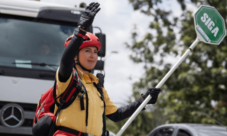 Persona con casco rojo, guantes y uniforme amarillo sostiene una paleta de señalización con la palabra “SIGA” mientras levanta una mano.