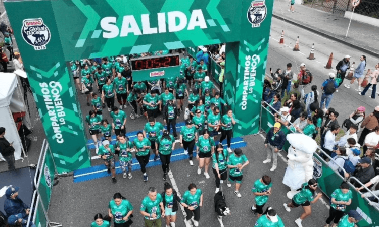 Cientos de participantes cruzan el arco de salida de la Bimbo Global Race 2026 sobre una vía urbana. El pórtico verde muestra el texto “SALIDA”, los logotipos de Bimbo Global Race 2025 y la frase “COMPARTIMOS LO BUENO”. A los lados hay espectadores detrás de vallas, mientras corredores con camisetas verdes avanzan por la línea de partida.