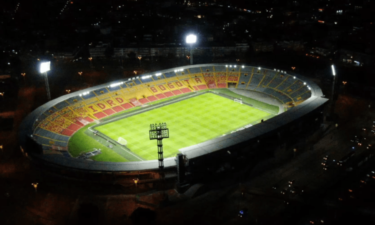 Vista nocturna del estadio El Campín iluminado durante un partido de fútbol, con las tribunas visibles alrededor del terreno de juego y las luminarias encendidas sobre la cancha.
