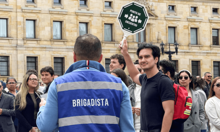 Grupo de personas reunidas en una plaza frente a un edificio institucional. En primer plano, una persona con chaleco azul que dice “BRIGADISTA” observa a otra que sostiene una señal octagonal con el texto “PUNTO DE ENCUENTRO”.