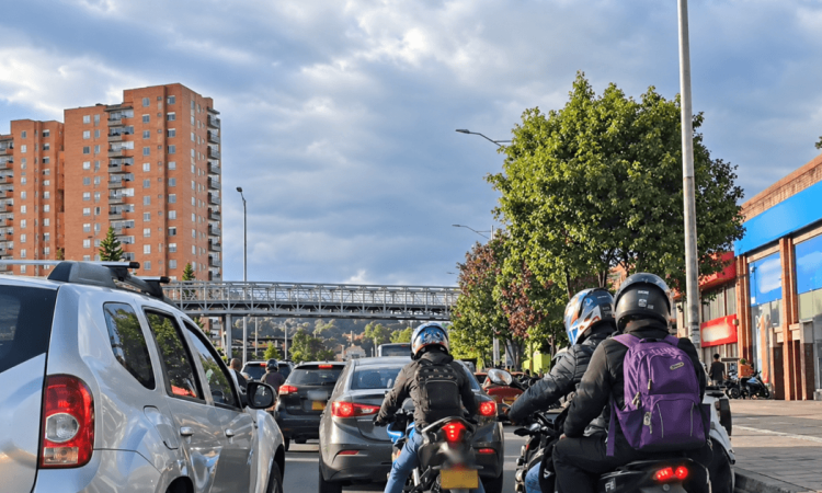 Motociclistas avanzan entre vehículos en una vía de Bogotá durante el día. A un costado de la carretera se observan árboles y, al fondo, edificios residenciales bajo un cielo parcialmente nublado.