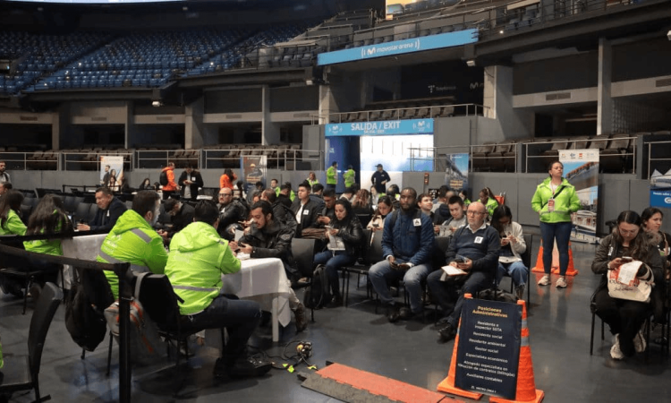 Personas sentadas en filas dentro de un recinto ferial mientras participan en una jornada de empleo. En primer plano hay mesas de atención y personas con chalecos institucionales; al fondo se observan estands, pantallas informativas y asistentes reunidos.