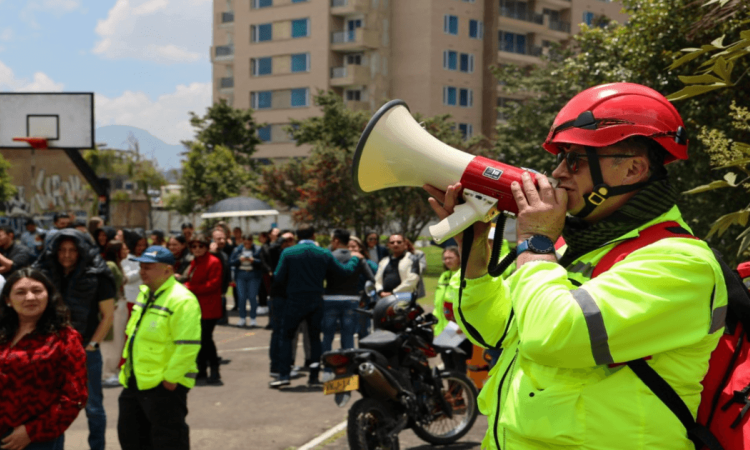 Persona con casco rojo y chaqueta reflectiva usa un megáfono para orientar a un grupo de ciudadanos durante un ejercicio de simulación de emergencia por sismo y otras emergencias en un espacio urbano de Bogotá.