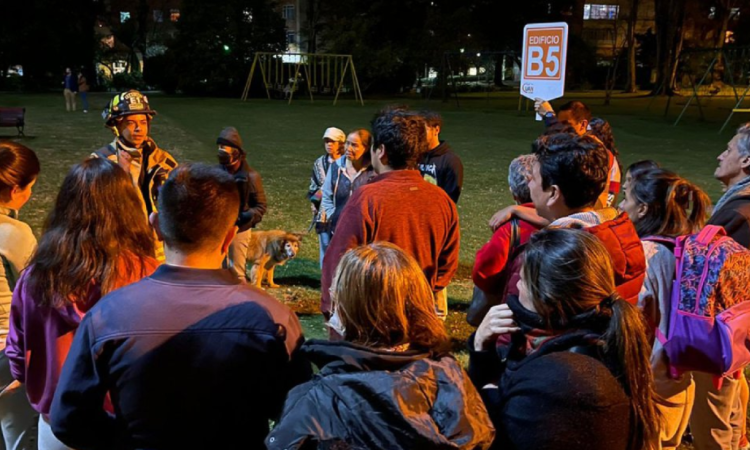 Grupo de personas reunidas en un parque durante una jornada nocturna de preparación para simulacro. Un integrante del cuerpo de bomberos brinda indicaciones mientras una persona sostiene un letrero con la identificación "Edificio B5".