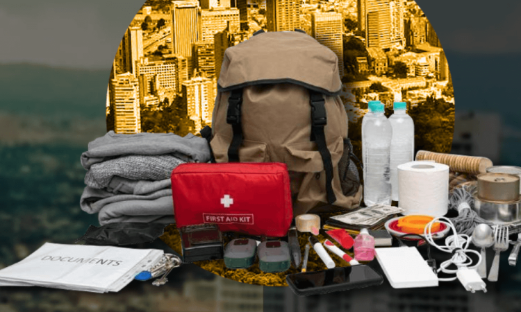 Kit de emergencias sobre una mesa con una mochila gris, botiquín rojo, linterna, pilas, radio portátil, alimentos no perecederos, botellas de agua y otros elementos recomendados para atender una emergencia.