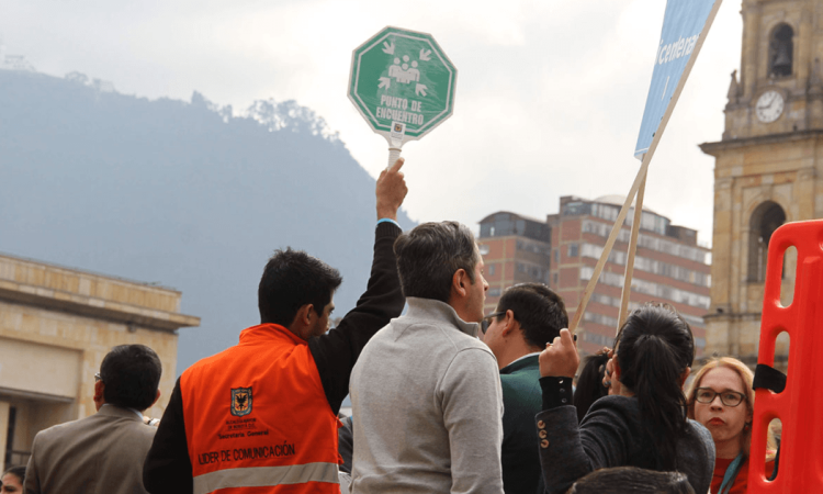 Personal de logística y participantes reunidos en una plaza del centro de Bogotá durante un simulacro. Una persona sostiene en alto una señal verde de “Punto de encuentro”, mientras alrededor se observan varios asistentes. Imagen de referencia para el Simulacro Bogotá 2026.