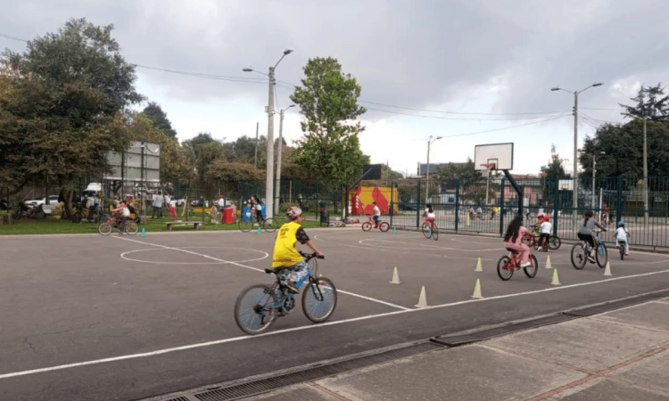 Persona con casco amarillo monta una bicicleta entre conos de señalización instalados en una pista durante Vacaciones Recreativas del IDRD con actividades de formación ciclista al aire libre.
