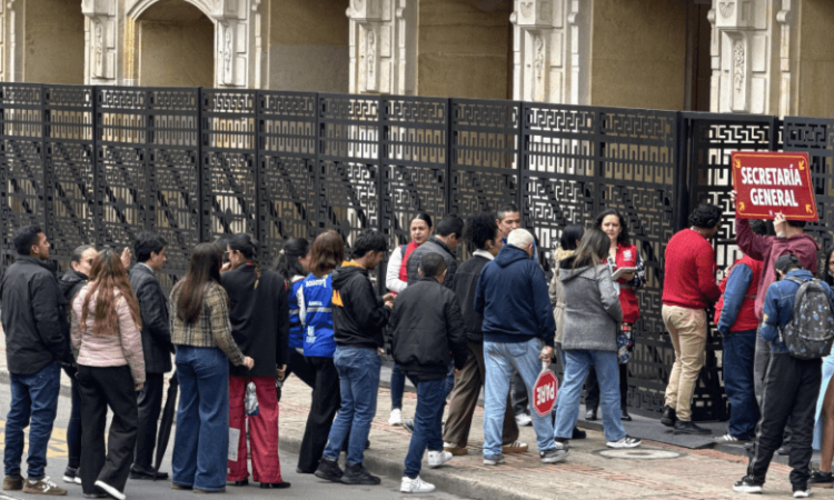 Grupo de personas reunidas sobre un andén junto a una vía, frente a una reja metálica negra. Varias personas llevan maletas, morrales y bolsos, mientras permanecen agrupadas cerca de la entrada de un edificio de fachada clara.