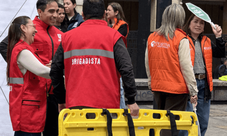 Grupo numeroso de personas concentradas alrededor de una señal verde con el texto “PUNTO DE ENCUENTRO”. La actividad se desarrolla en una plaza pública rodeada de edificaciones y estructuras urbanas